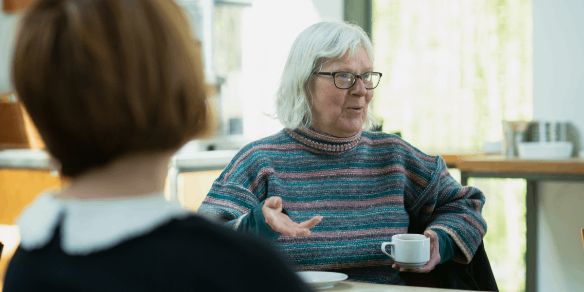 Two women talking. The older woman is holding a white cup.