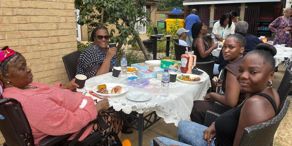 Four people are sat around a table in the garden smiling and sharing food