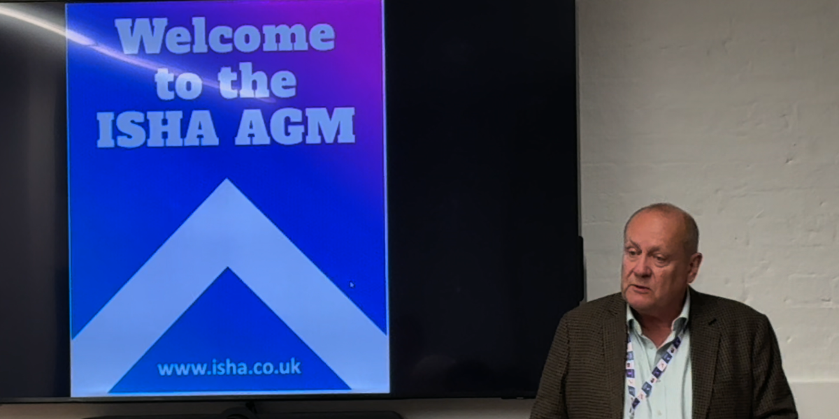 Bob Heapy, is speaking, and standing in front of a board that has a sign which reads Welcome to the ISHA AGM