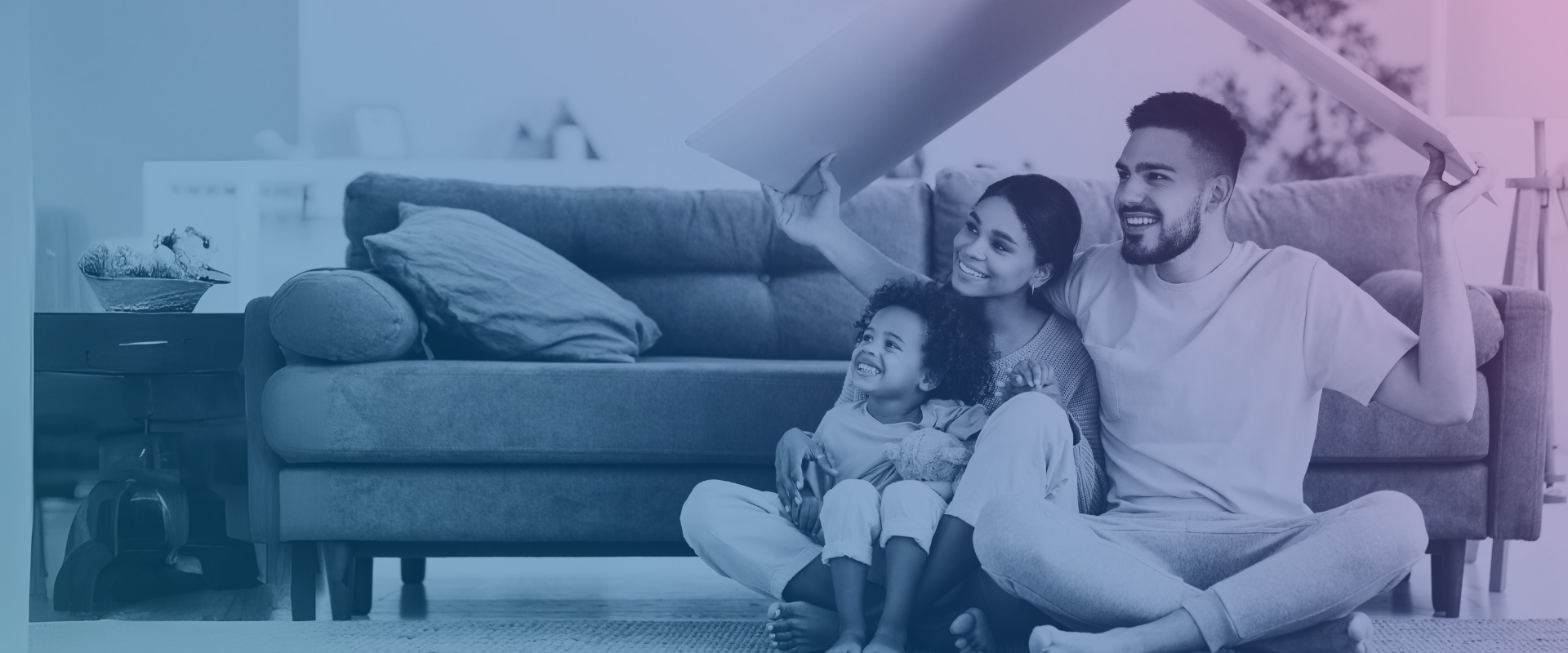 A smiling family sitting in front of a sofa, holding a cardboard above their head in the shape of a roof.